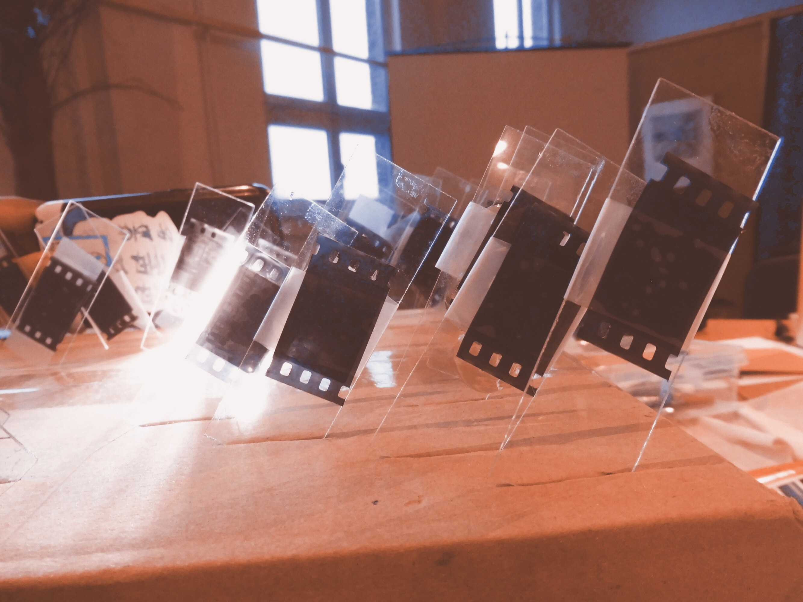 A cardboard box is photographed at an angle. There are slits cut in the upper face. Photographic negatives are threaded into the slits. The light from a portable lamp illuminates the negatives on the left. A laptop computer, a window, and a projection screen can be glimpsed in the background.