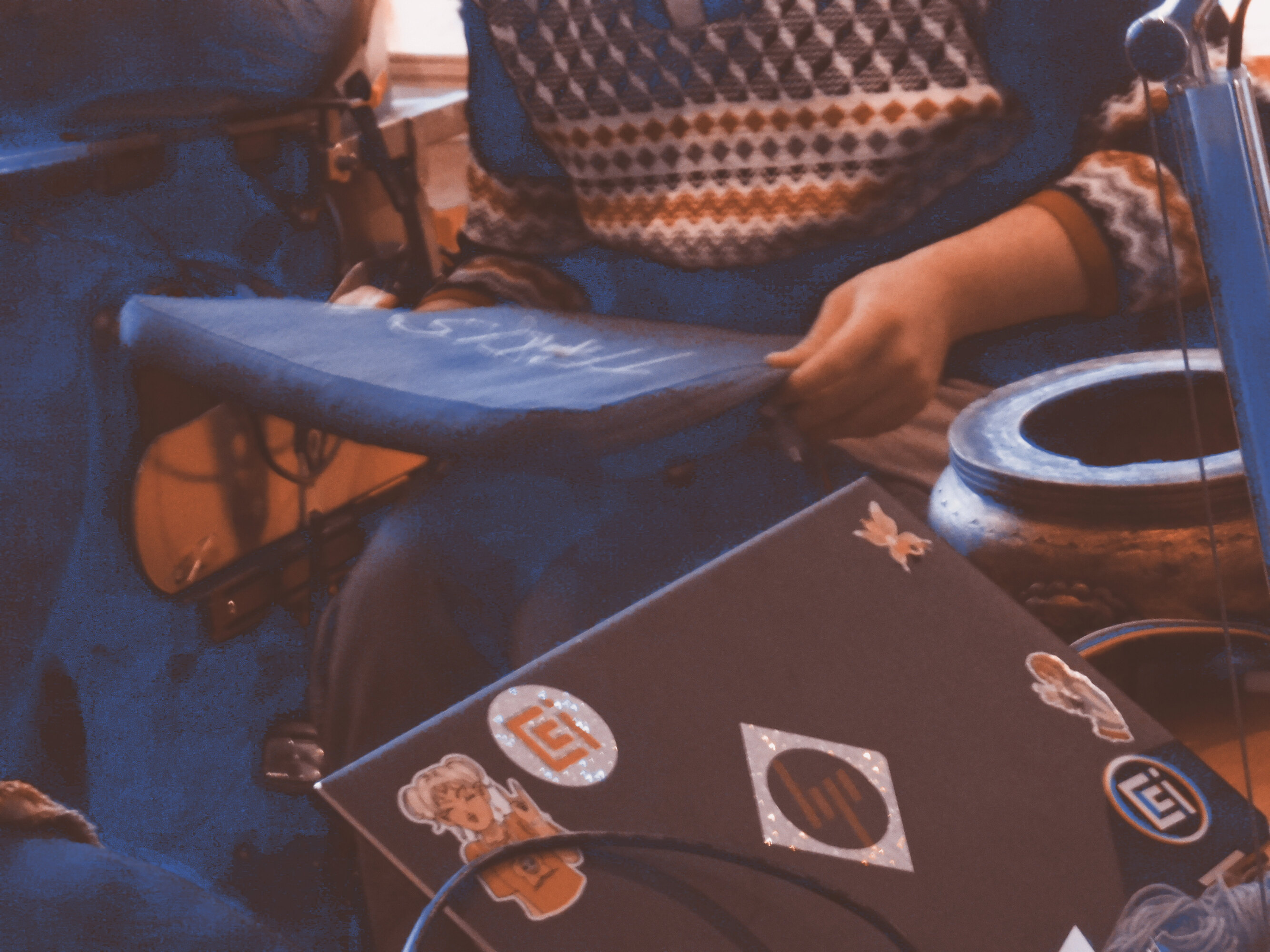 A person is holding a flat grey pillow with two hands. On the pillow is sewn the word *Trust* with white thread. Another person is holding on their lap a black portable computer covered with stickers.