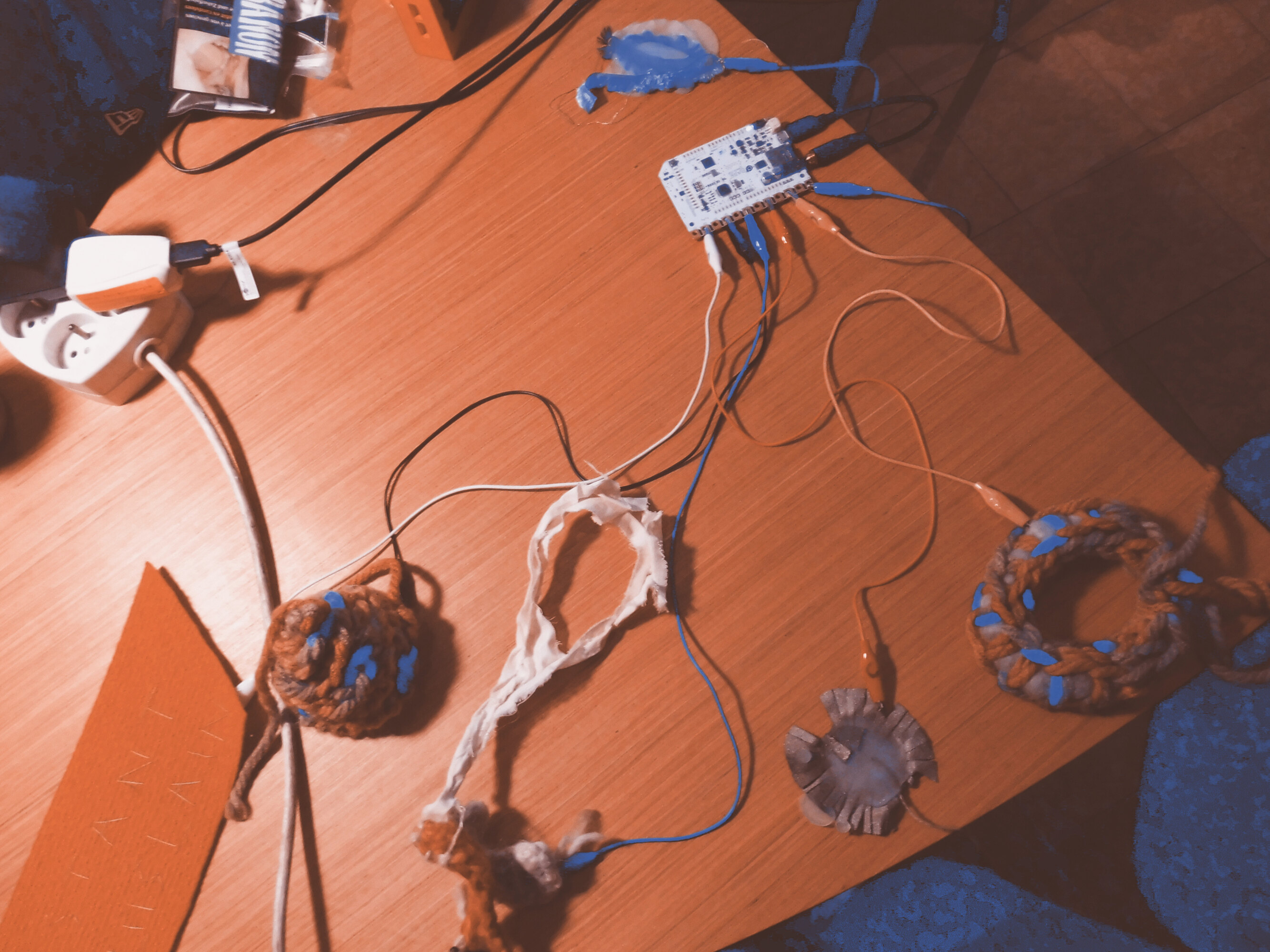 A photo taken from above of an installation resting on a wooden table. The installation consists of colorful cables, electrical circuits, a socket, and crocheted and fabric objects.