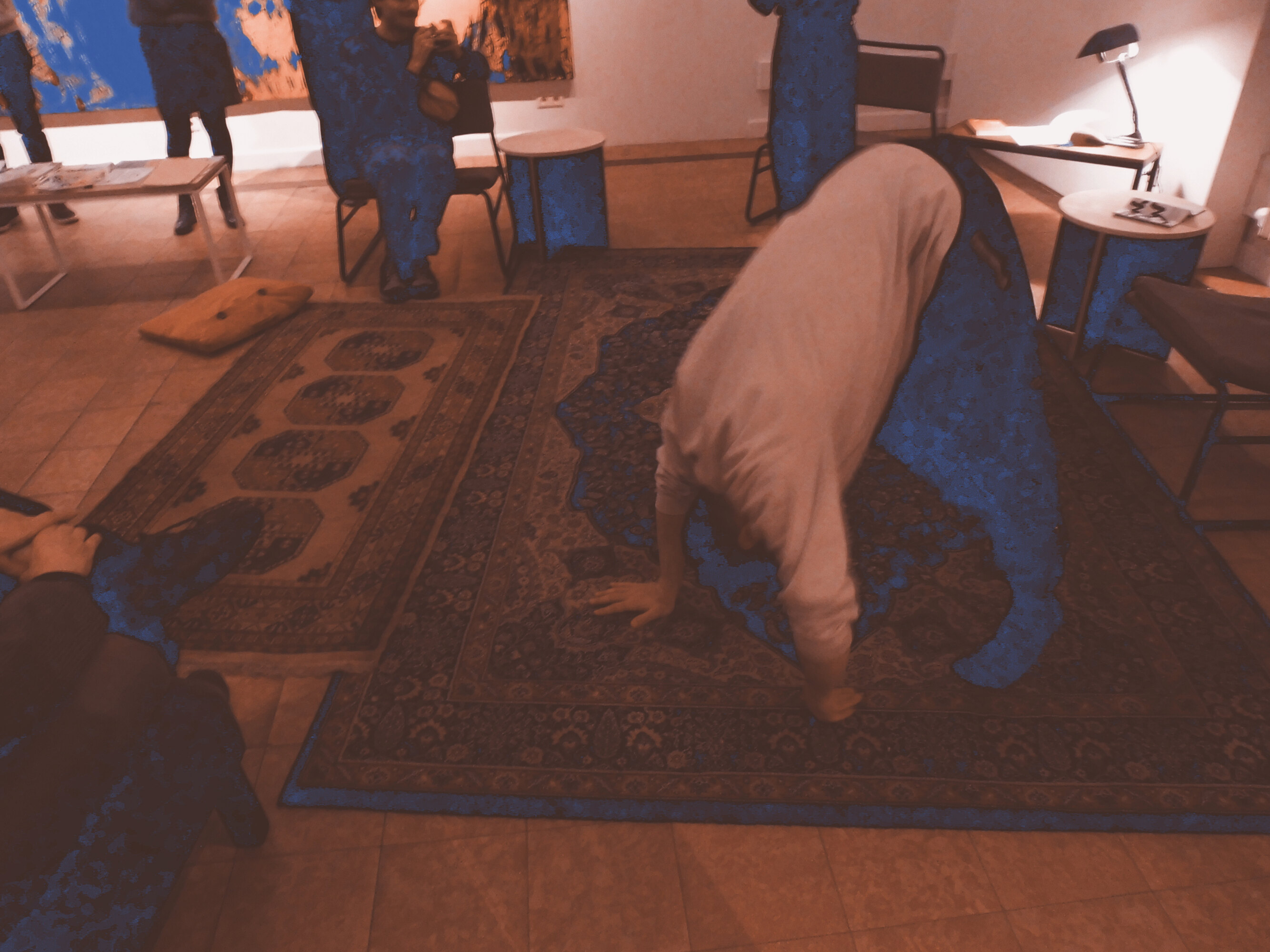 This is a picture of a dance choreography in a series of four. A dancer dressed with dark trousers and a white, long-sleeves T-shirt is bending to the floor. There are two carpets on the floor, one big and one small, with colorfoul patterns. People are standing or sitting all around the dancer. There are also small tables and armchairs and a lamp.