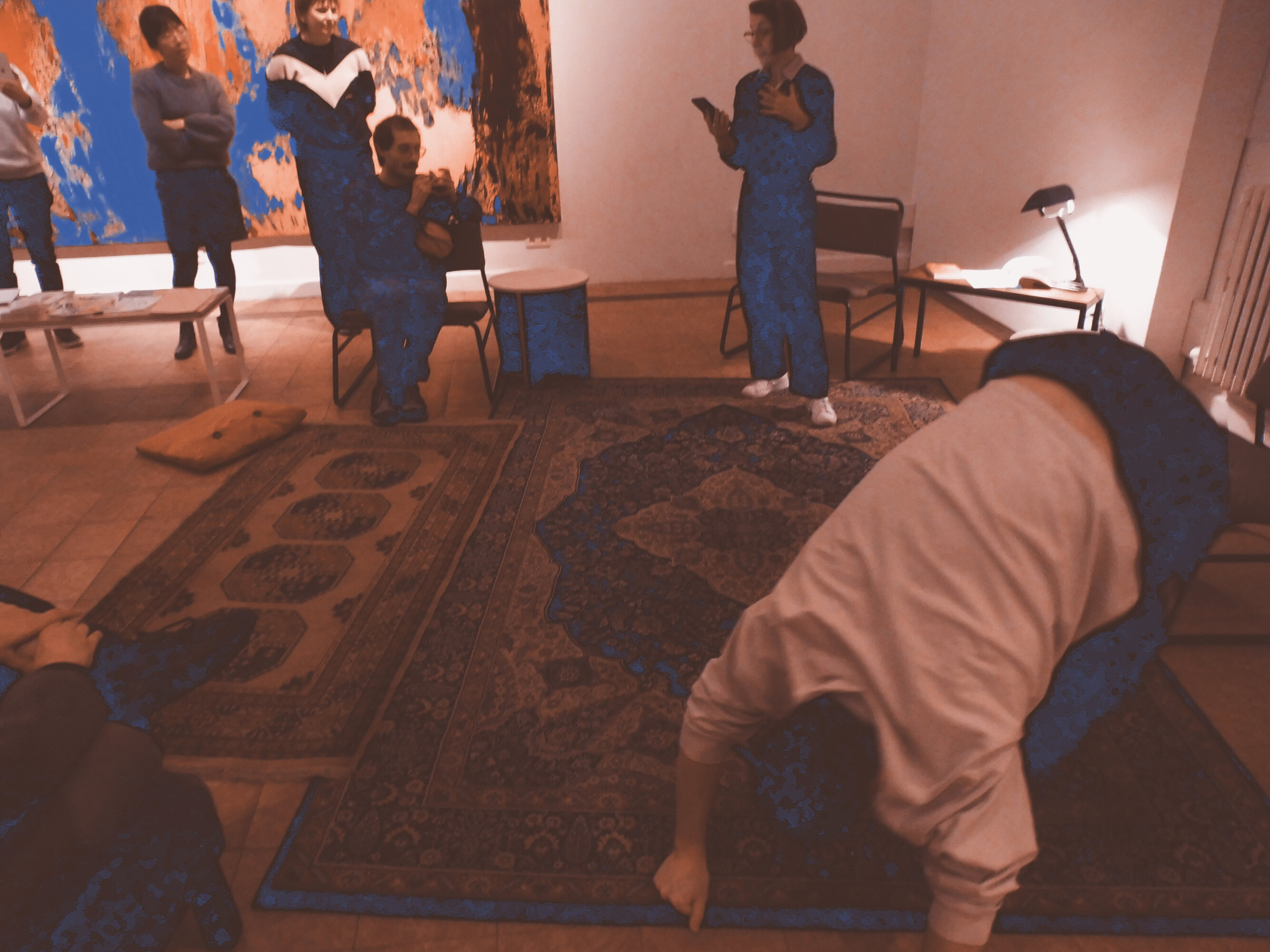 This is a picture of a dance choreography in a series of four. A dancer dressed with dark trousers and a white, long-sleeves T-shirt is bending to the floor, leaning on his left side.There are two carpets on the floor, one big and one small, with colorfoul patterns. Behind the dancer a person is standing. She is dressed in black with white shoes, she wears glasses and is reading on a telephone. People are standing or sitting all around the dancer. There are also small tables and armchairs and a lamp.