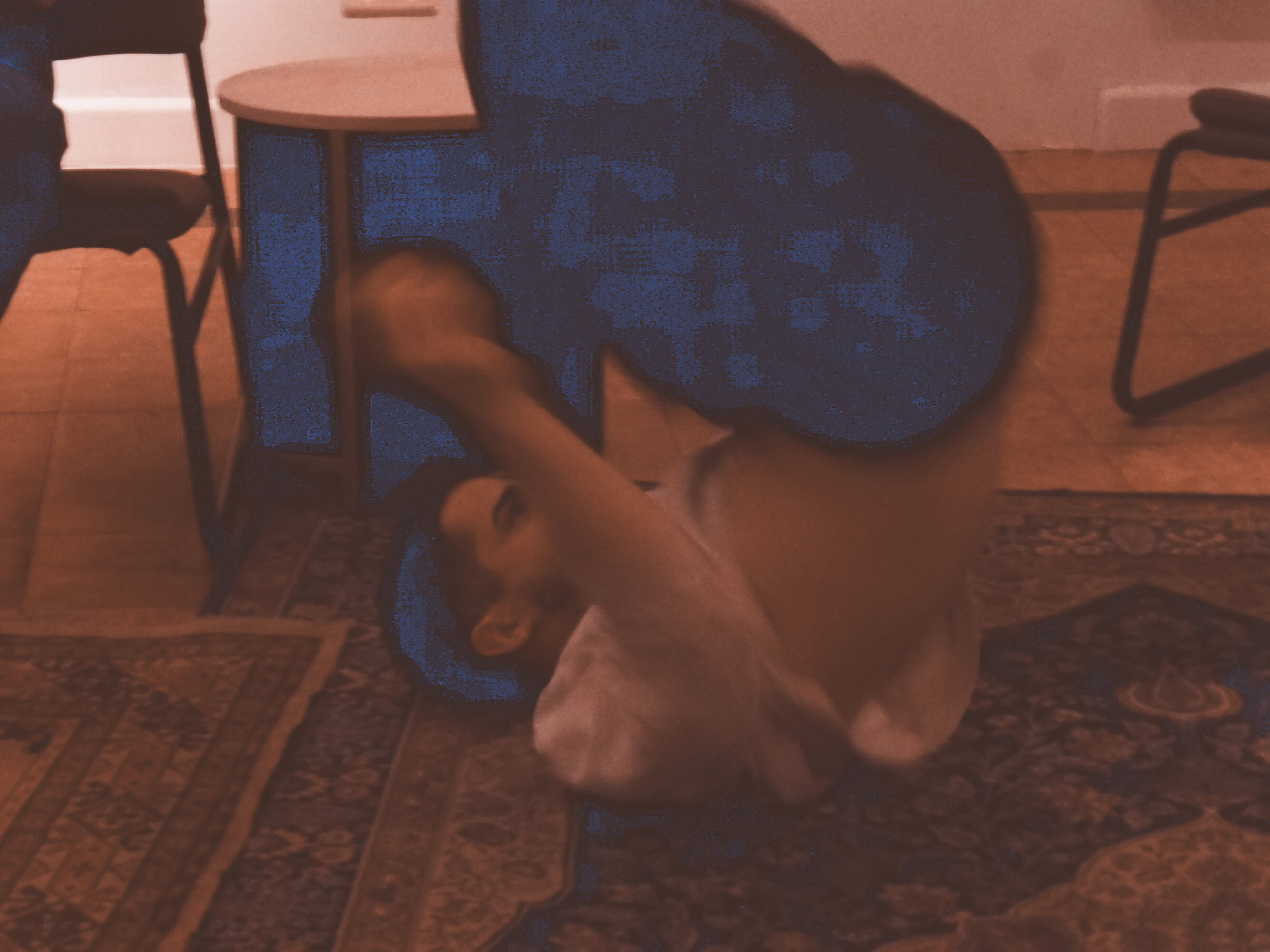 This is a picture of a dance choreography in a series of four. A close-up of a dancer dressed with dark trousers and a white, long-sleeves T-shirt while he is rolling with his legs up on a colorful carpet.