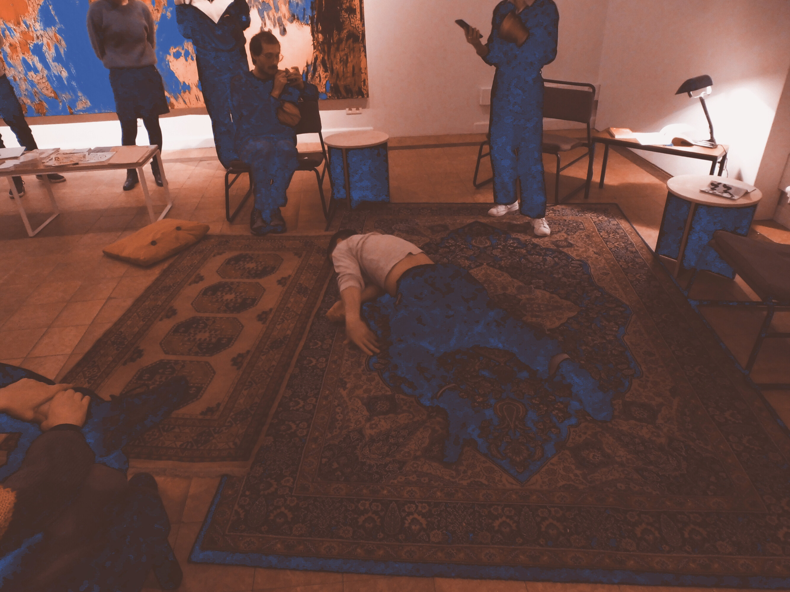 This is a picture of a dance choreography in a series of four. A dancer dressed with dark trousers and a white, long-sleeves T-shirt is lying on his belly on the floor. There are two carpets on the floor, one big and one small, with colorfoul patterns. People are standing or sitting all around the dancer. There are also small tables and armchairs and a lamp.