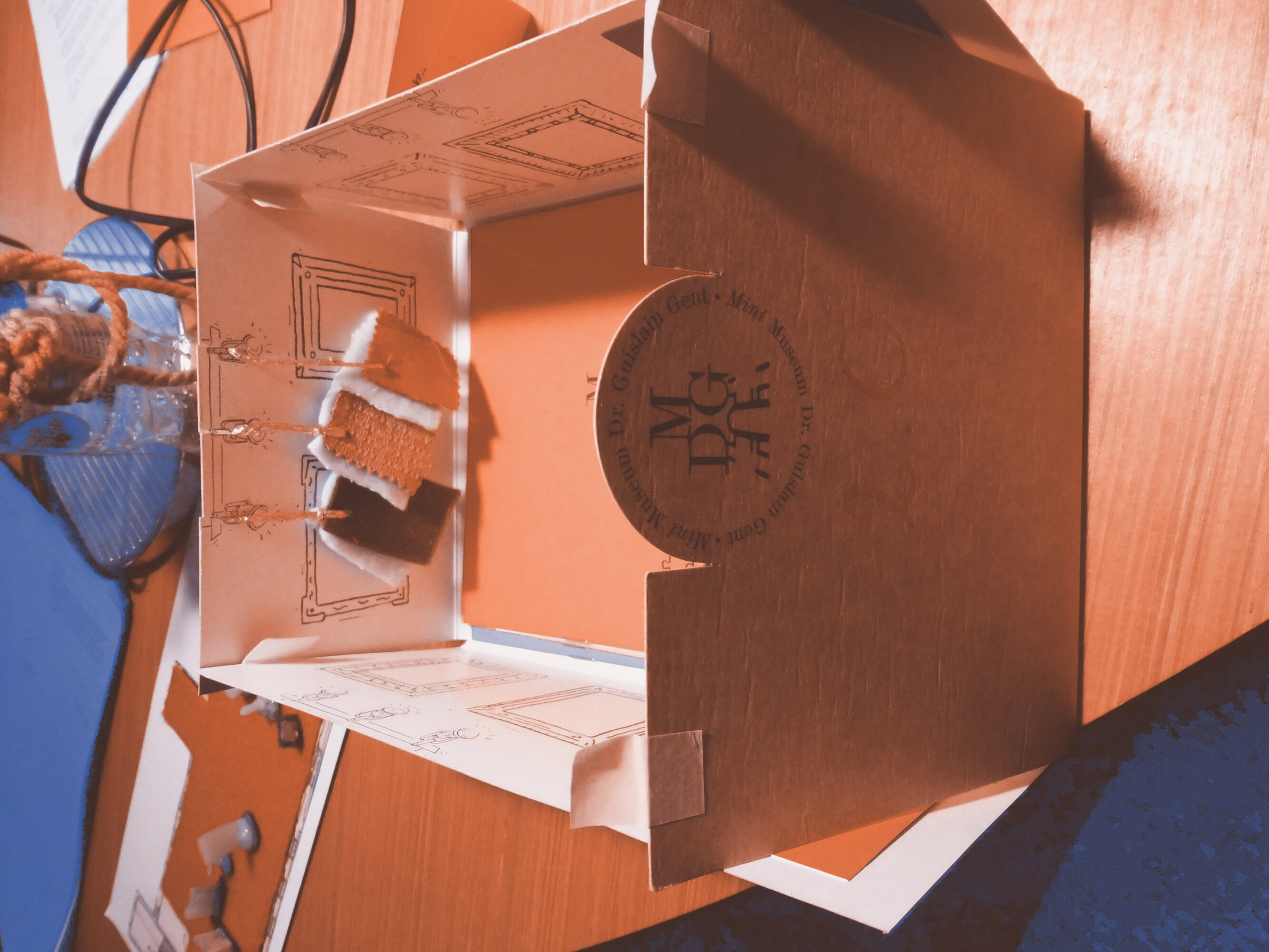 A cardboard box with the museum logo is on a table. The upper face is missing and you can see the inside. The inner part of the box is drawn in black and white like a museum room with paintings hanging on the walls. Three small squared cloths of different colors are attached with a golden cord to one inner side of the box.