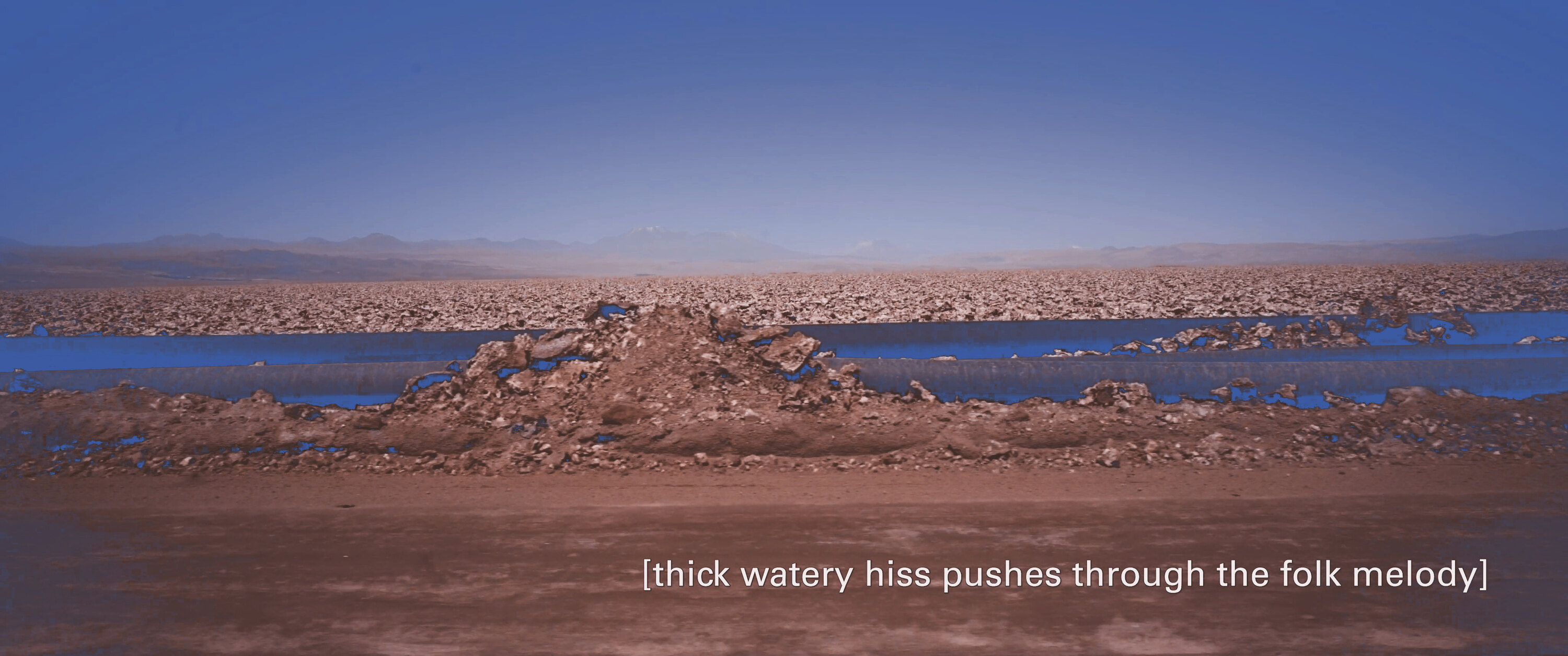 Screen capture from the movie: large conduits running along a road in the middle of a rocky desert, stretching back in the foggy horizon, with the caption “thick watery hiss pushes through the folk melody”